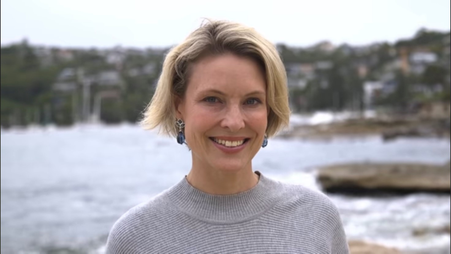A blonde woman looks at the camera in a coastal setting